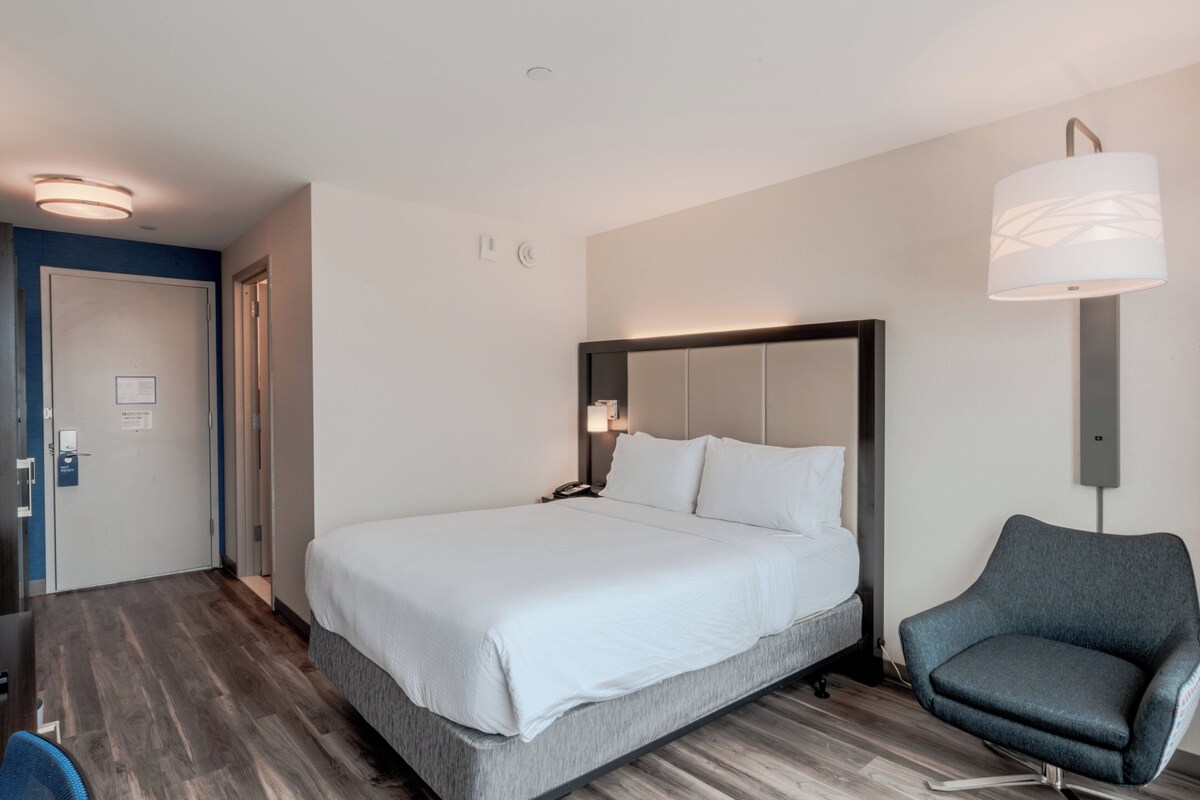 Una habitación de hotel con una cama grande, una mesita de noche, una silla y una lámpara de pared.