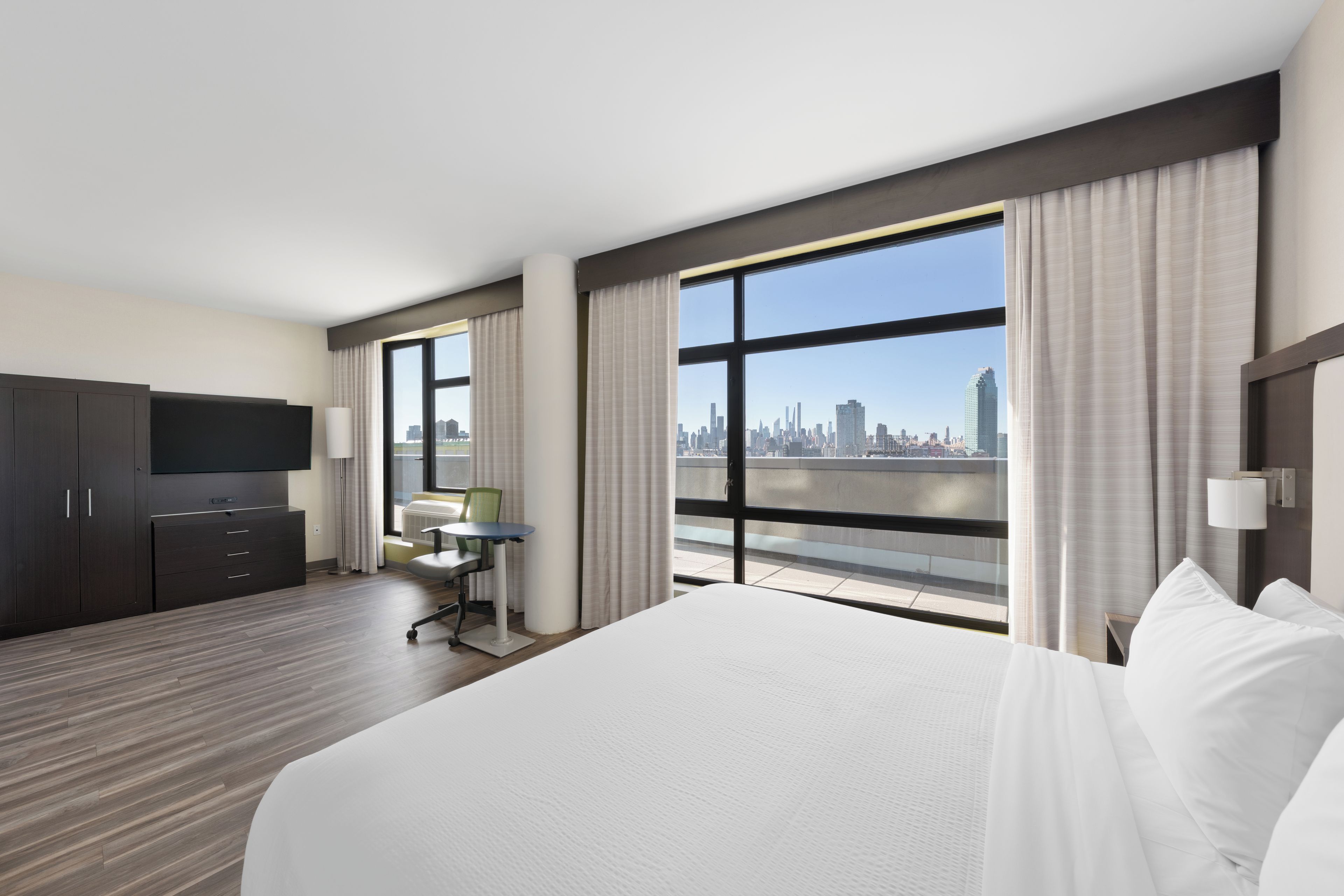 A hotel room with a large bed, a desk, a chair, a television, and a view of a city skyline.