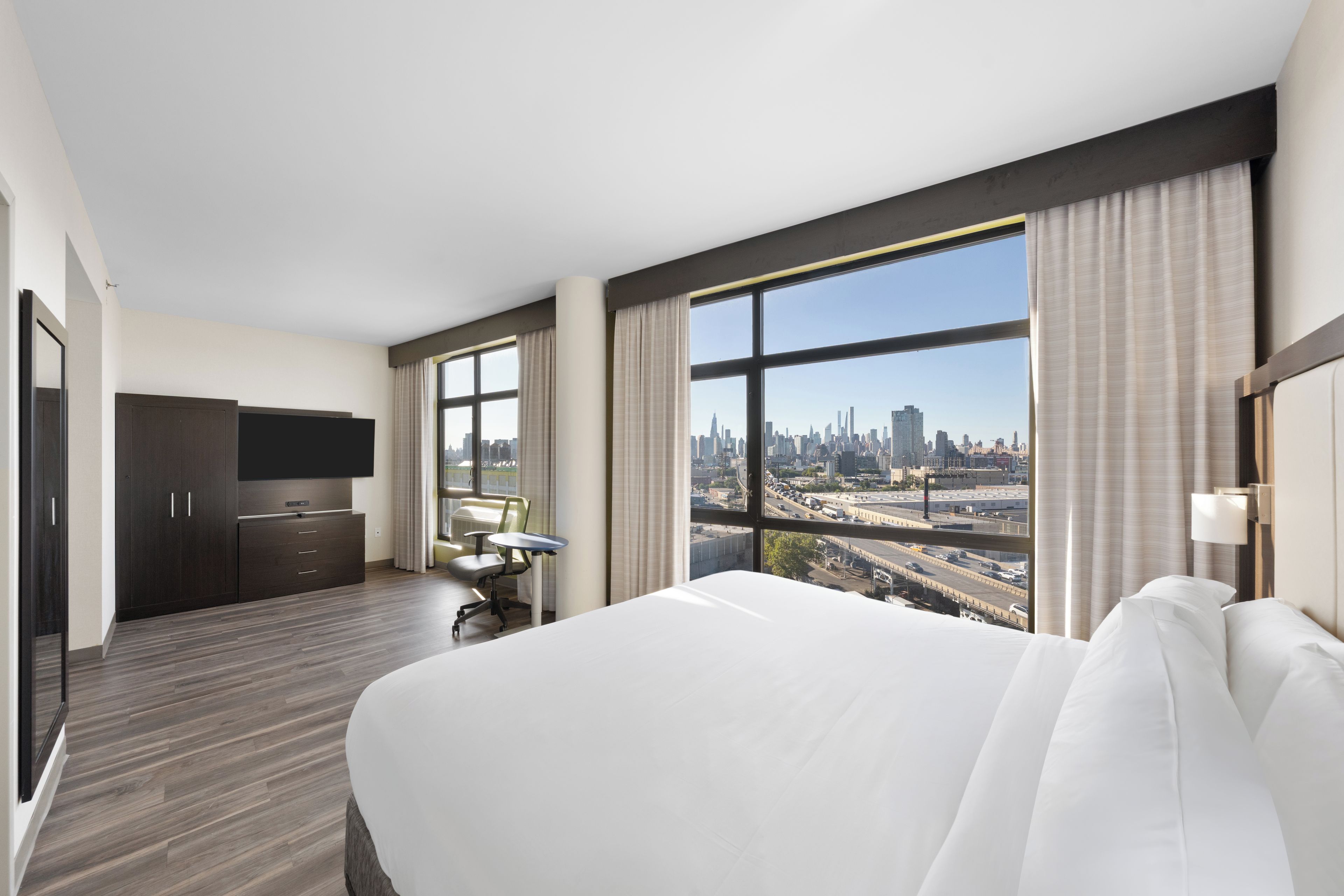A hotel room with a large bed, a desk, a chair, and a TV. There is a window with a city view.