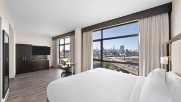 A hotel room with a large bed, a desk, a chair, and a TV. There is a window with a city view.