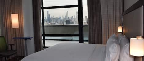 Habitación de hotel con una cama grande, dos lámparas de noche, una mesita y una silla. Se observa un horizonte urbano a través de la ventana.