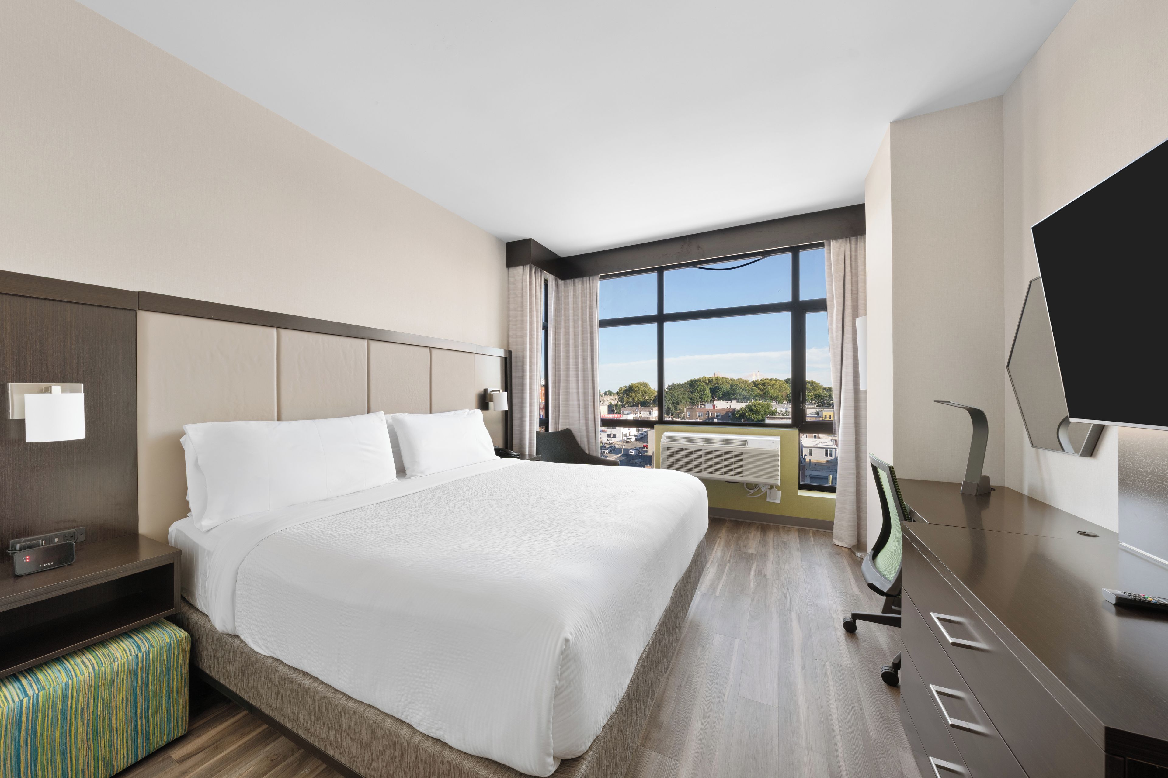 A hotel room with a large bed, a desk, a chair, a TV, and a window with a city view.