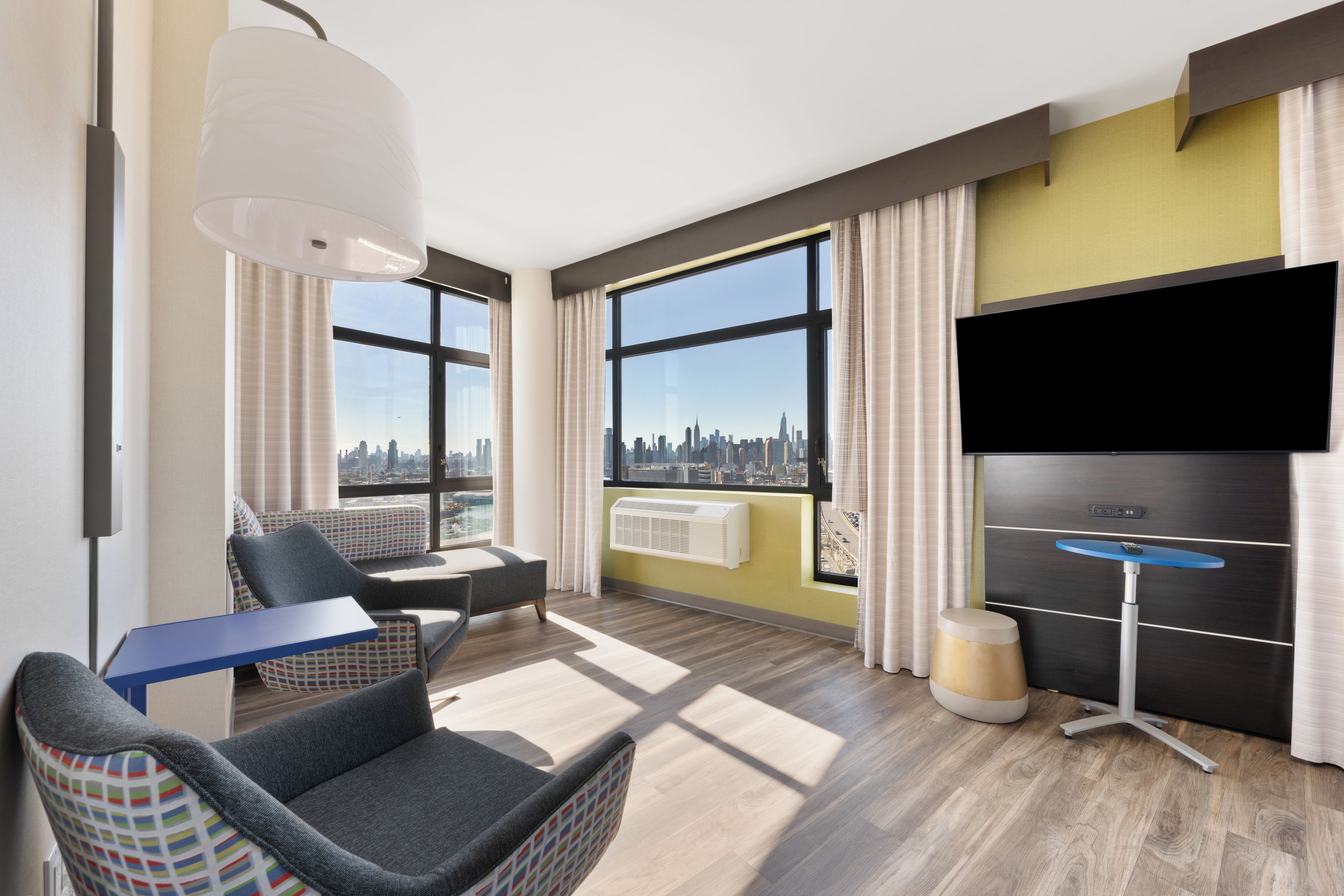 Een moderne hotelkamer met een groot raam, een flatscreen televisie, een bank, een klein tafeltje en een stoel.