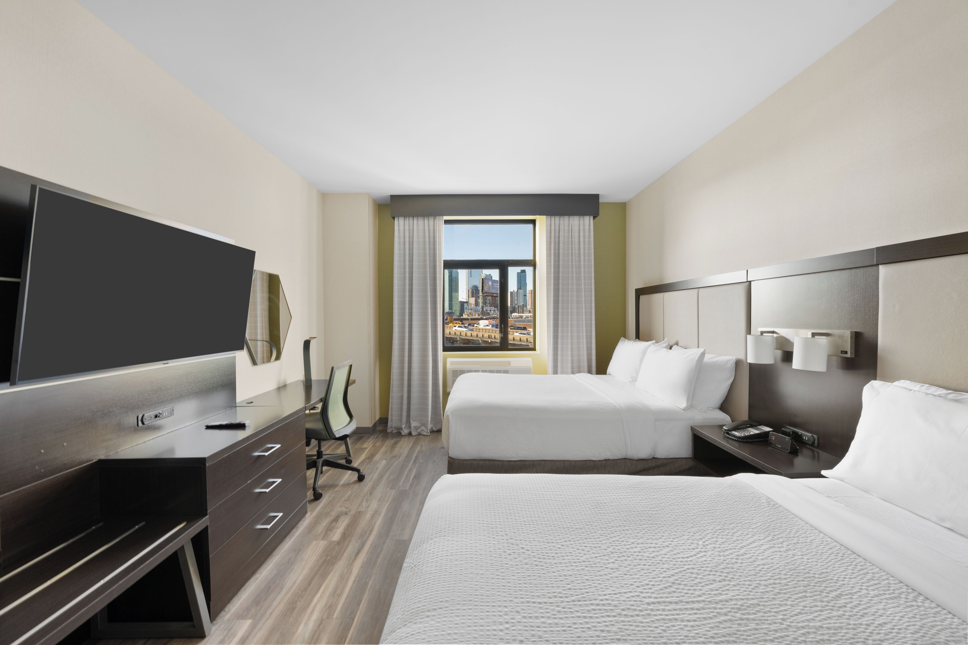 A hotel room with a large bed, a smaller bed, a desk with a TV, a chair, and a view of the cityscape through the window.