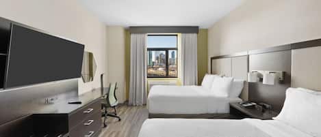 A hotel room with a large bed, a smaller bed, a desk with a TV, a chair, and a view of the cityscape through the window.