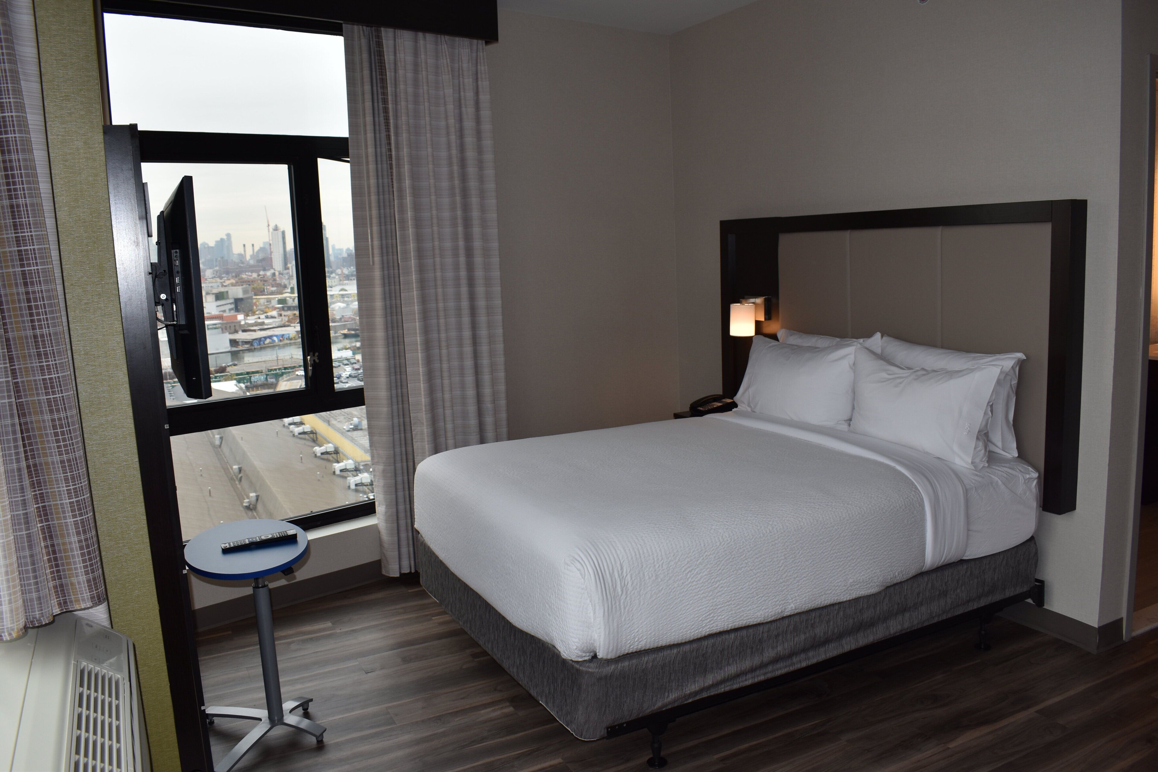 A hotel room with a large bed, a small table, a TV, and a window with a city view.
