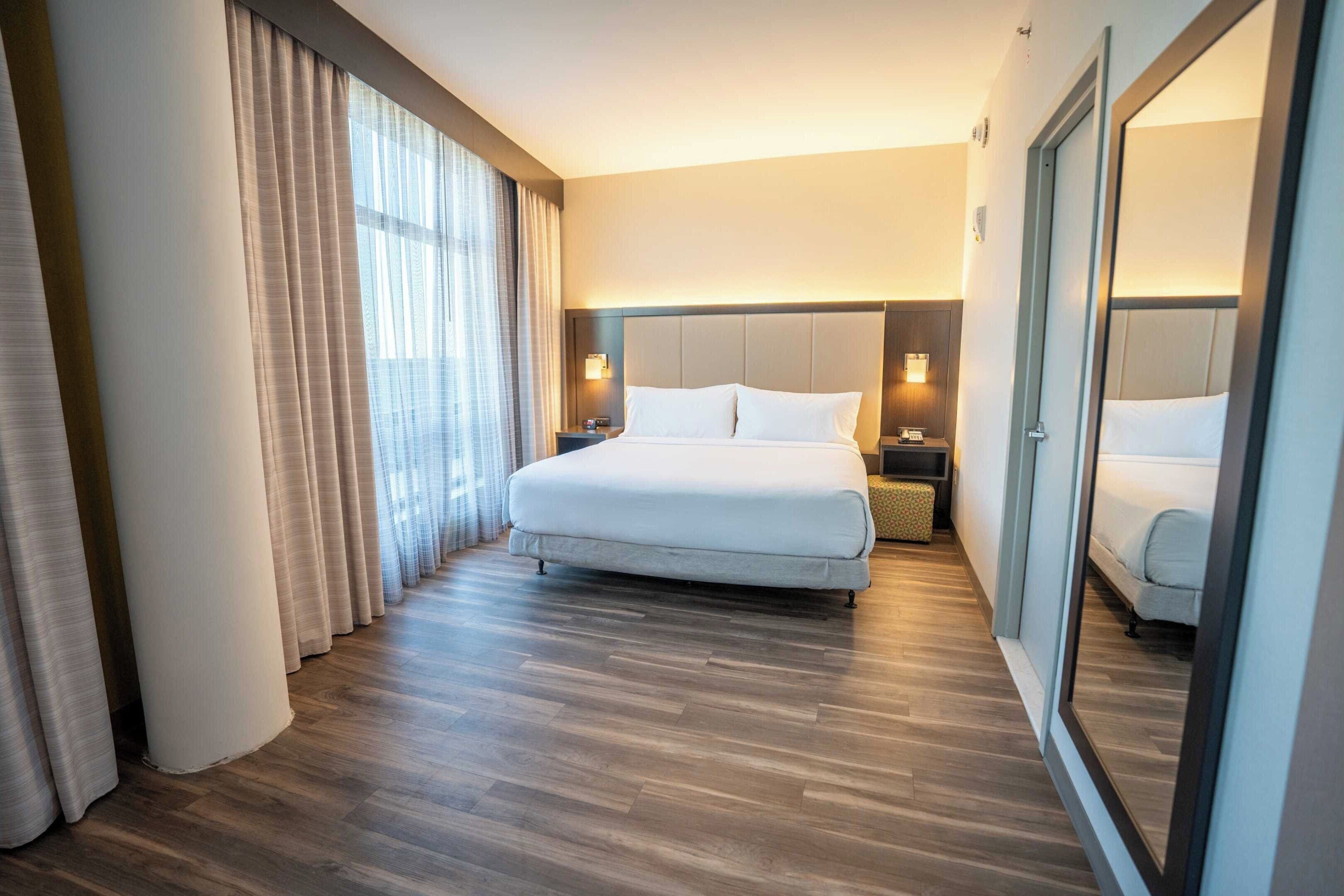 A hotel room with a large bed, wooden flooring, a nightstand with a lamp, and a large mirror.
