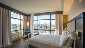 Habitación de hotel con una cama grande, un escritorio y vistas a la ciudad a través de amplios ventanales.