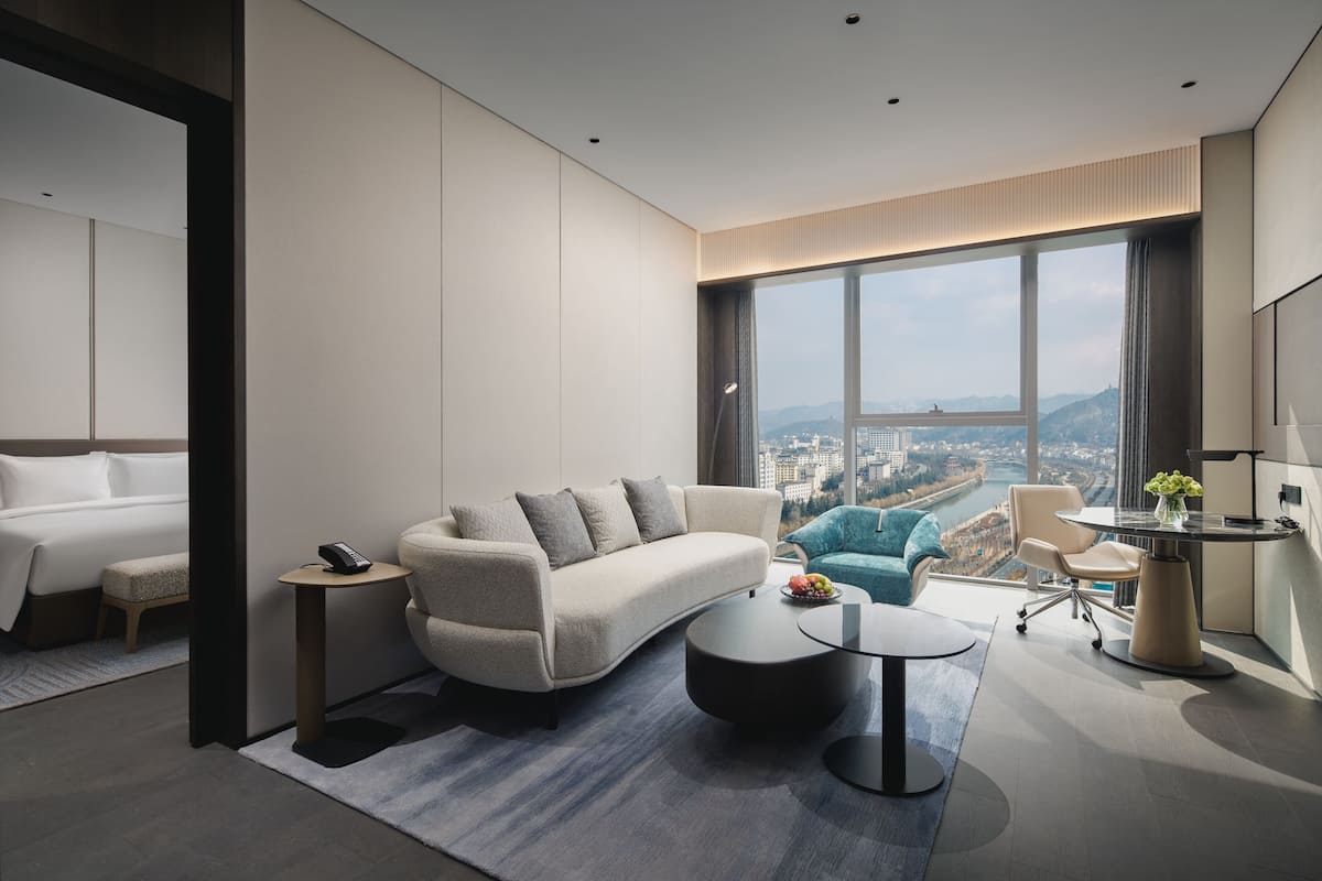 Una habitación de hotel moderna con un ventanal grande, un sofá, una mesita de café, un escritorio y una silla.