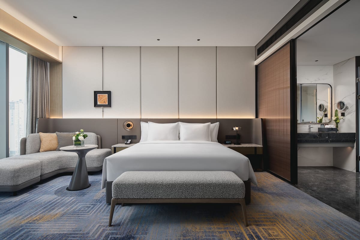 Una habitación de hotel moderna con una cama grande, un sofá, una mesita y un baño con espejo y lavabo.