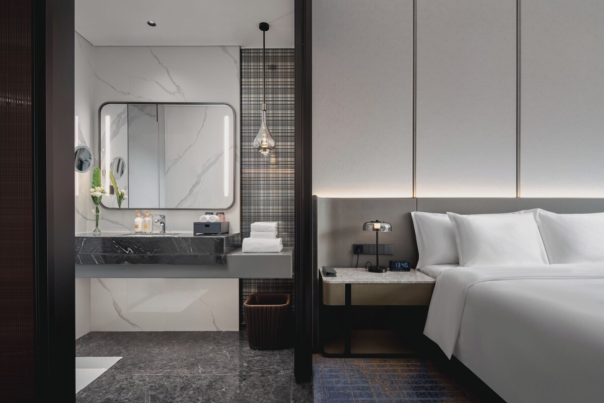Una habitación de hotel moderna con baño de mármol, cama con ropa blanca y mesita de noche con lámpara.