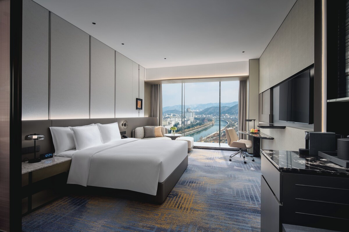 Habitación de hotel moderna con una cama grande, un escritorio, un televisor y vistas a la ciudad y montañas.
