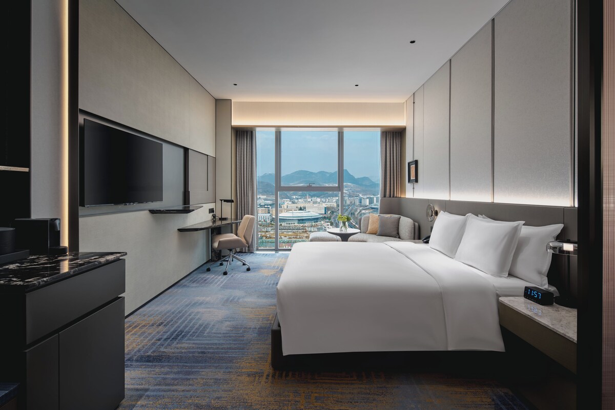 Una habitación de hotel moderna con una cama grande, un televisor de pantalla plana montado en la pared, un escritorio con silla y vistas a un paisaje urbano a través de amplios ventanales.