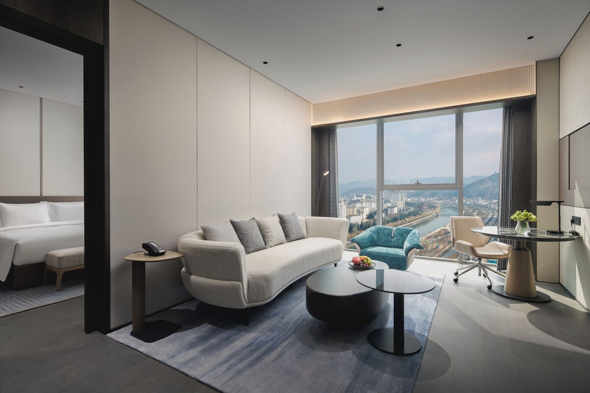 Una habitación de hotel moderna con un ventanal grande, un sofá, una mesa de centro y un escritorio con una lámpara.