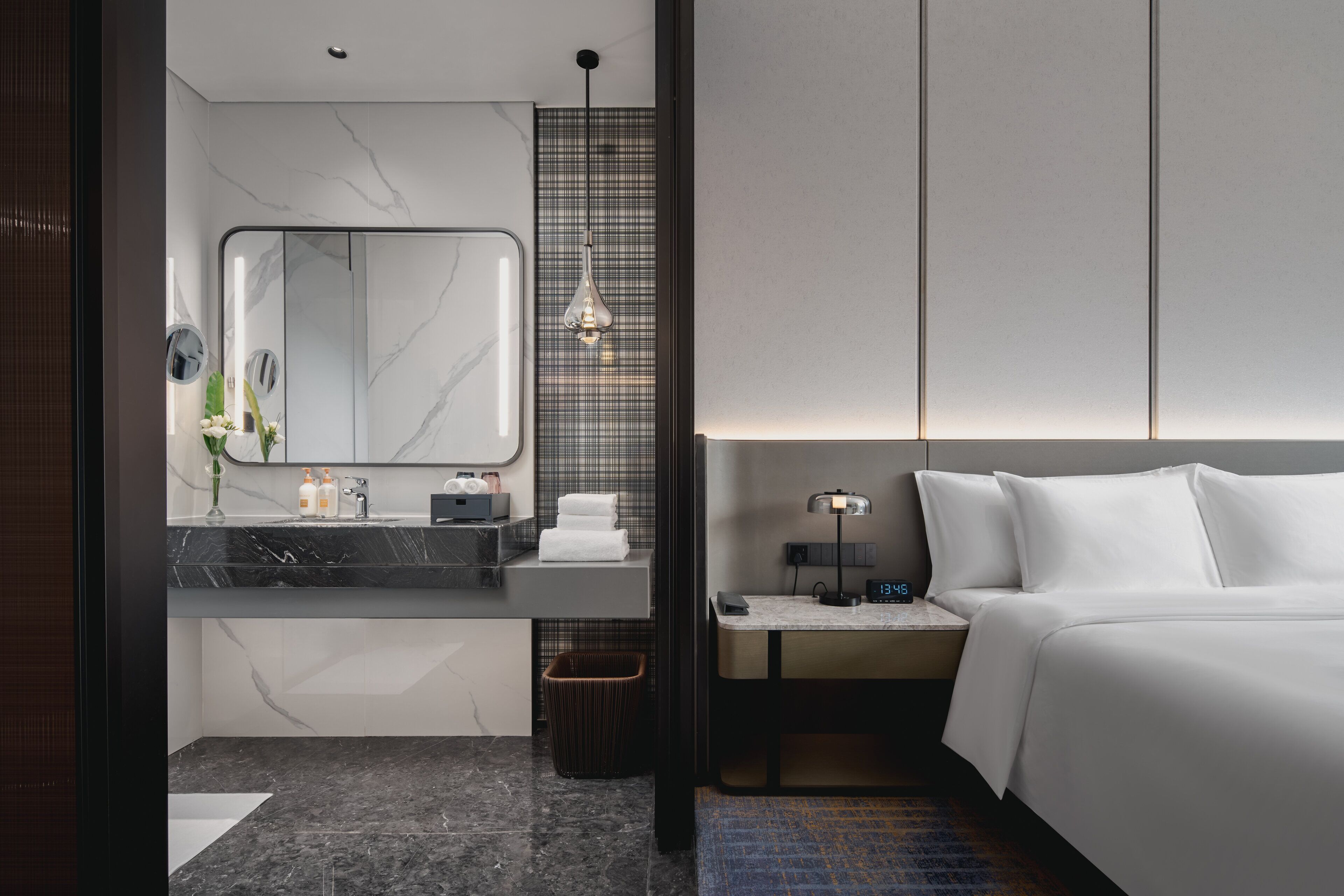 Habitación de hotel moderna con baño de mármol, una mesita de noche con lámpara y una cama con ropa blanca.