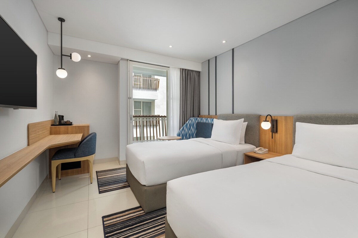 A modern hotel room with two beds, a desk, a chair, a TV, and a balcony with a view.