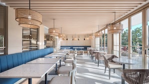 A modern restaurant with a long bench seating area, tables, wicker chairs, and large windows offering a view.