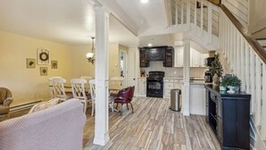 Un vaste salon doté d’un haut plafond, un coin repas avec des chaises blanches et une cuisine avec des meubles sombres et un plancher en bois.