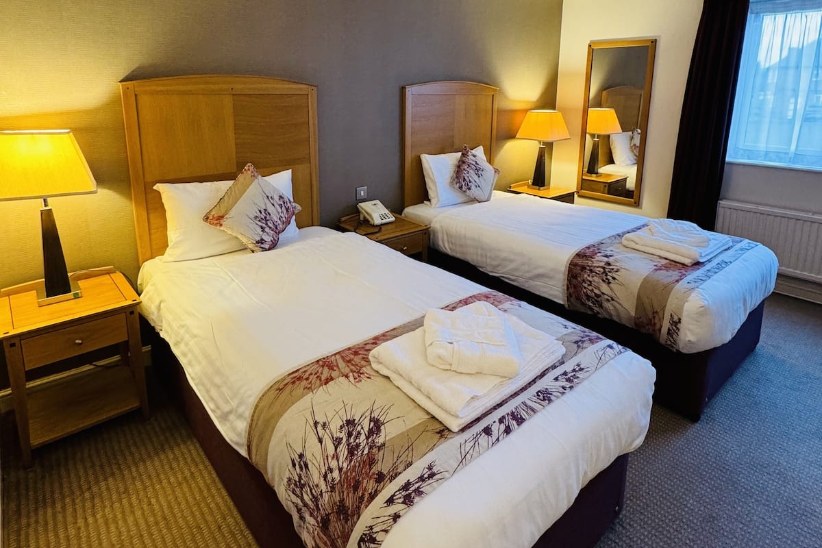 Une chambre d’hôtel avec deux lits, chacun doté d’une tête de lit en bois, de tables de chevet avec des lampes et d’une grande fenêtre avec des rideaux.