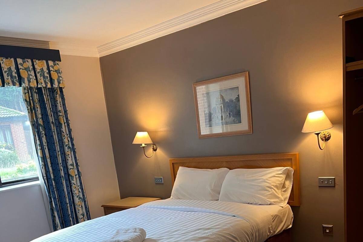 Un lit bien fait, avec du linge de lit blanc et une serviette pliée, une tête de lit en bois, deux lampes fixées au mur et un tableau encadré au-dessus du lit.