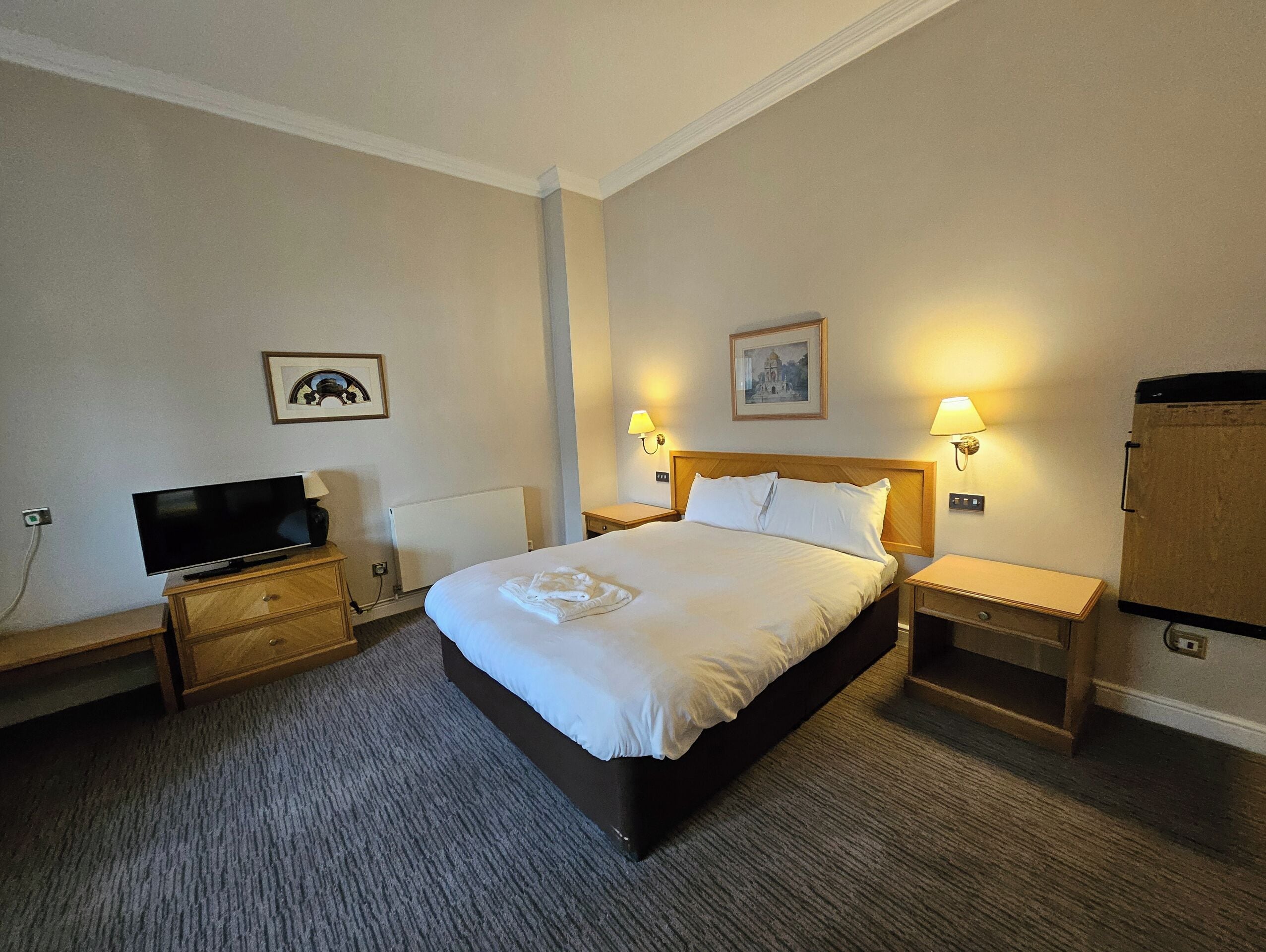 Habitación de hotel con una cama grande, un televisor de pantalla plana, dos lámparas de noche, un cuadro enmarcado en la pared y un mueble de madera.