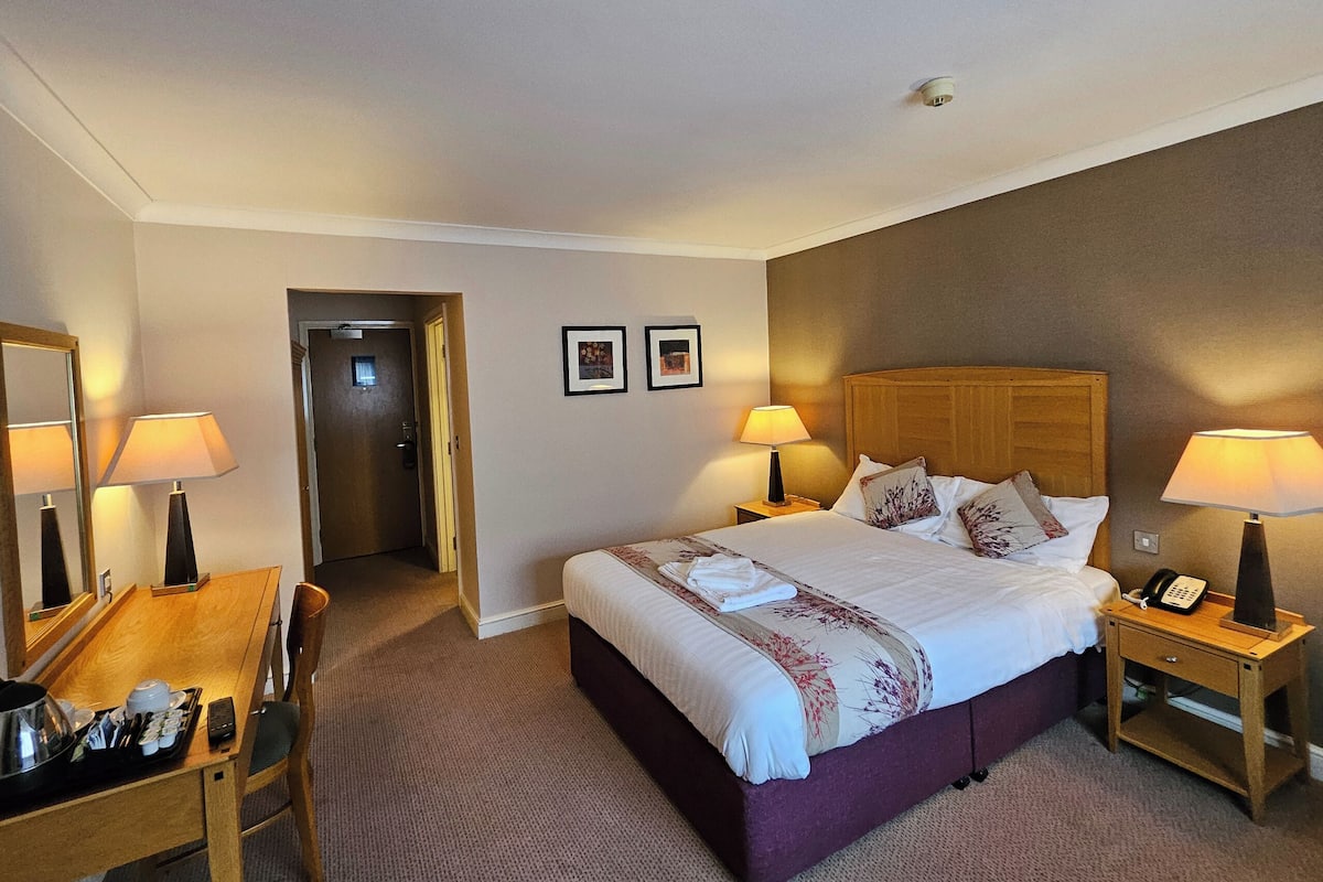 Une chambre d’hôtel comprenant un lit, des lampes de chevet, un bureau avec un téléphone et une porte donnant sur une autre pièce.