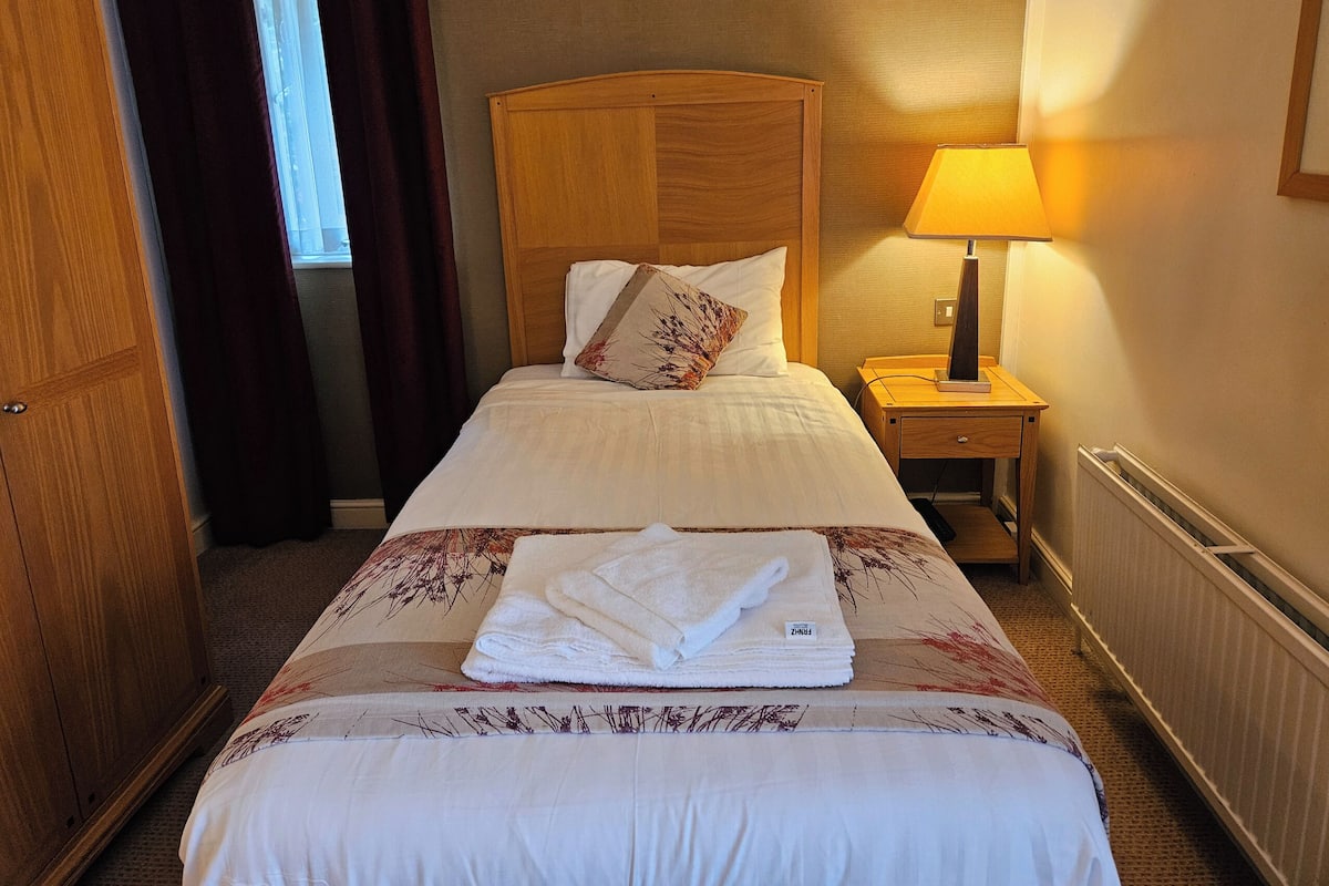 Une chambre d’hôtel avec un lit, une table de chevet avec une lampe, une armoire en bois et une fenêtre avec des rideaux.