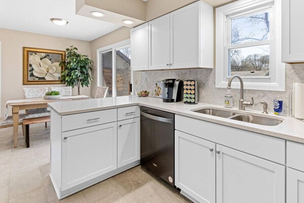 Una cocina moderna con armarios blancos, lavavajillas, cafetera y un lavabo.