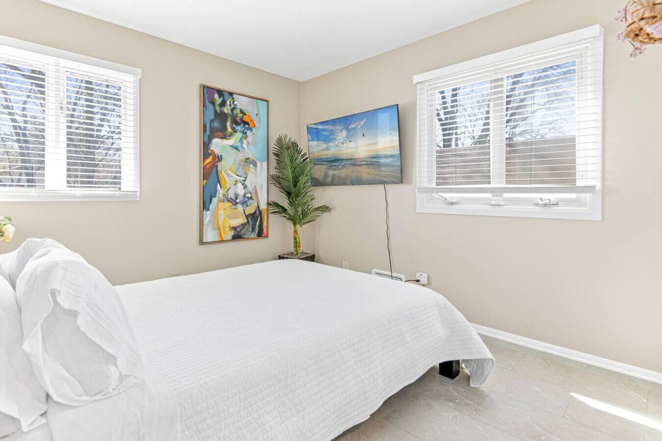 A bedroom with a bed, two windows with blinds, a potted plant, and two paintings on the wall.