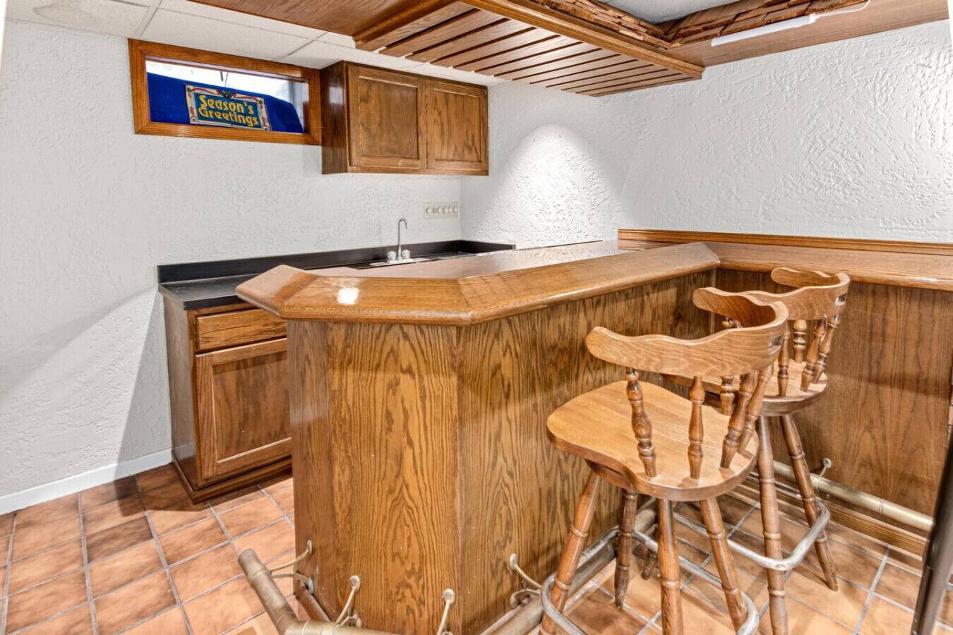 A kitchen with wooden cabinets, a bar area with stools, and a sign above the window reading "Season's Greetings".