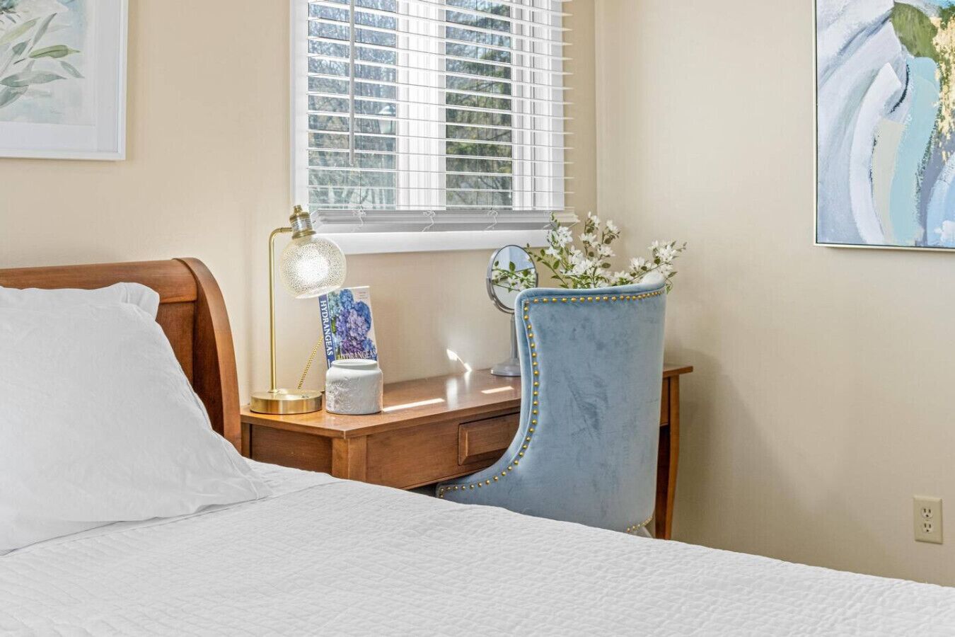 A neatly made bed with white linens, a wooden nightstand with a lamp, a blue upholstered chair, and a window with blinds.