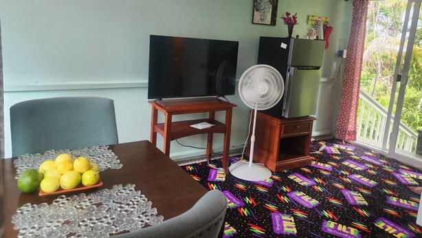 Quarto com mesa de jantar, cadeiras, televisão de tela plana, ventilador, geladeira e varanda com vista para vegetação.