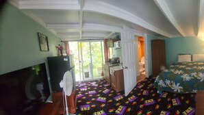 Quarto de hotel com cama, escrivaninha, ventilador e janela com vista para o verde.