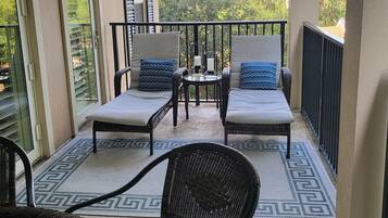 Un balcone con due poltrone imbottite e un tavolo, un tappeto decorato e vista su un edificio e alberi.