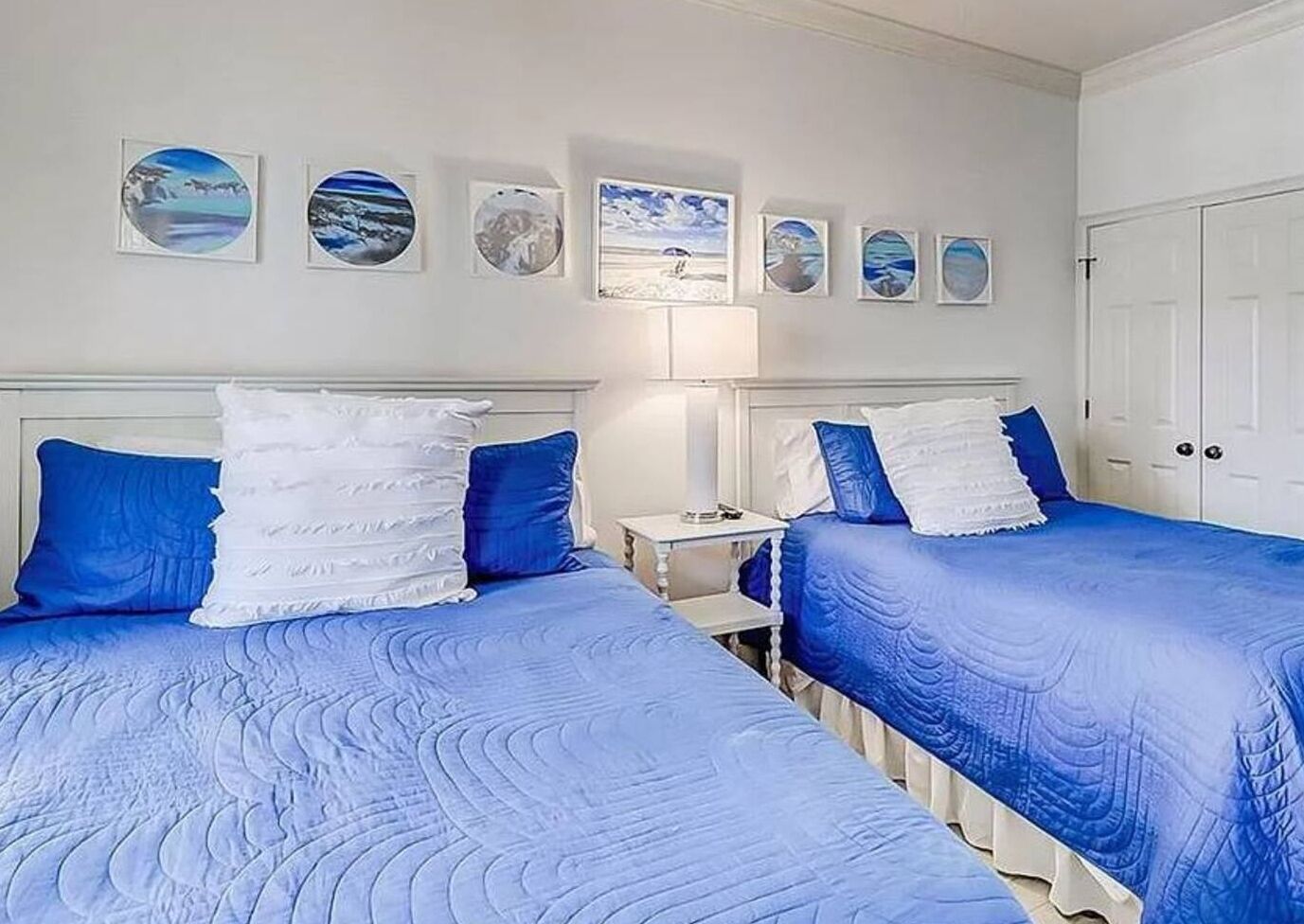 Una camera con due letti, ognuno con lenzuola blu e cuscini bianchi. Sopra i letti, ci sono quadri appesi al muro.