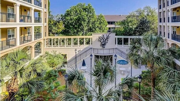 A courtyard with a central fountain, surrounded by palm trees and a large tree in the background. - Coligny Beach, Pools, & Shops- Few Steps Away (Hilton Head Island)