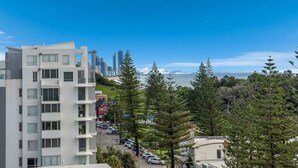 Un moderno edificio de apartamentos con varios pisos y balcones, rodeado de vegetación exuberante y una vista despejada del horizonte urbano y el océano.