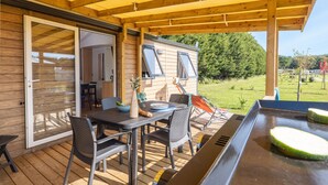 Una terraza de madera con una mesa, sillas, una parrilla y un ventanal corredero que da a una habitación.
