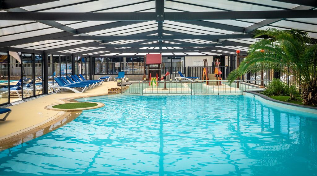 An indoor swimming pool with blue water, surrounded by lounge chairs and a covered area with a roof.