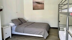 A single bed with a grey bedspread, a bedside table with a lamp, a bunk bed to the right, and a poster on the wall.