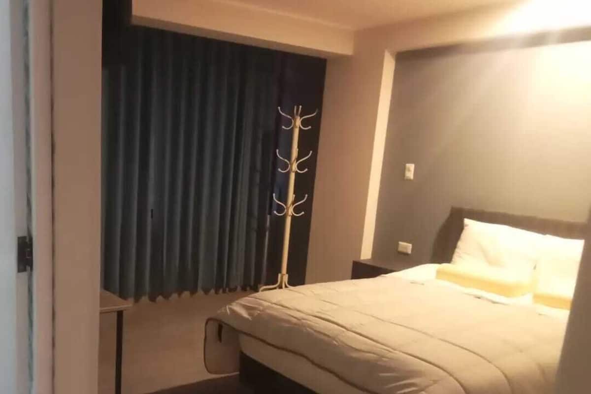 A bedroom with a bed, a coat rack, a window with curtains, and a nightstand.