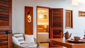 A cozy room with a wicker sofa, a wooden table with a teapot and cups, and a bed with a warm light. - Villa Nanui Jeri (Jijoca de Jericoacoara)