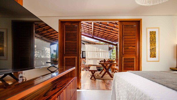 A bedroom with a bed, a wooden desk, and a view of an outdoor area with a table and chairs. - Villa Nanui Jeri (Jijoca de Jericoacoara)