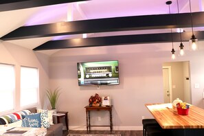 A modern living room with a television mounted on the wall, a wooden dining table, and a cozy seating area with a sofa and cushions. - Stylish 2BR Bungalow | Near Airport & Downtown DSM, Newly remodled and close to downtown Des Moines (Des Moines)
