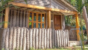 A thatched-roof structure with a wooden door and windows, surrounded by lush greenery. - Kings Villa Resort (Canacona)