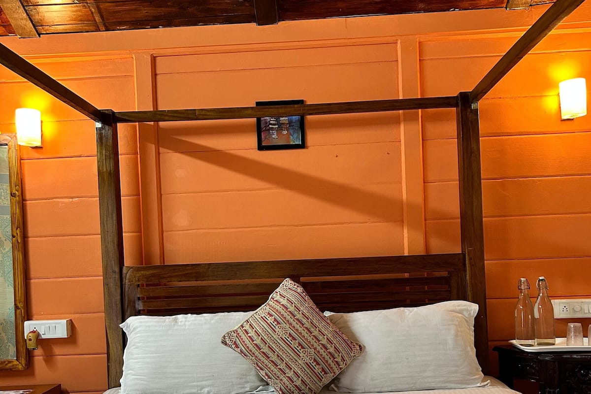 A bed with white linens, a wooden headboard, and a patterned pillow. There are two bottles and a glass on the nightstand beside the bed.
