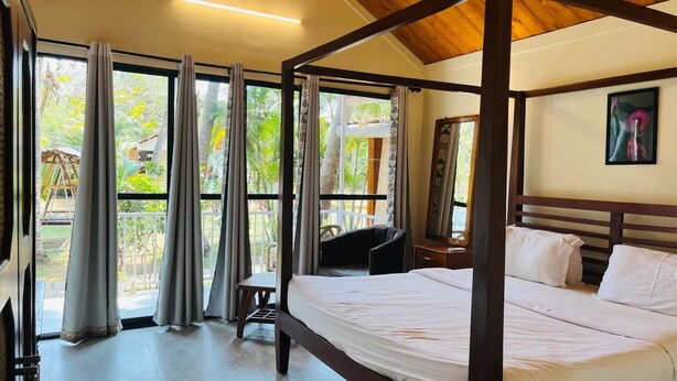 A bedroom with a large bed, a chair, a desk, and a window with curtains. - Kings Villa Resort (Canacona)