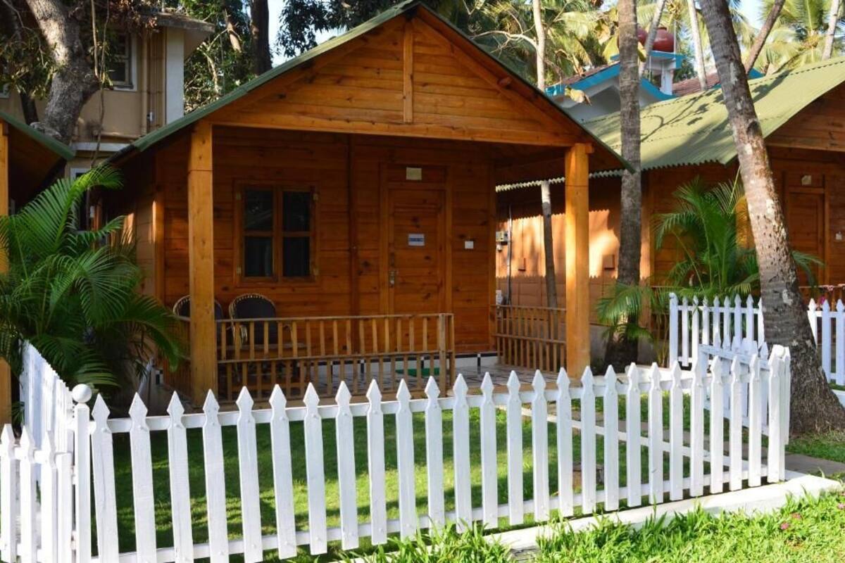 A wooden cabin with a porch, surrounded by a white picket fence, palm trees, and a small garden.