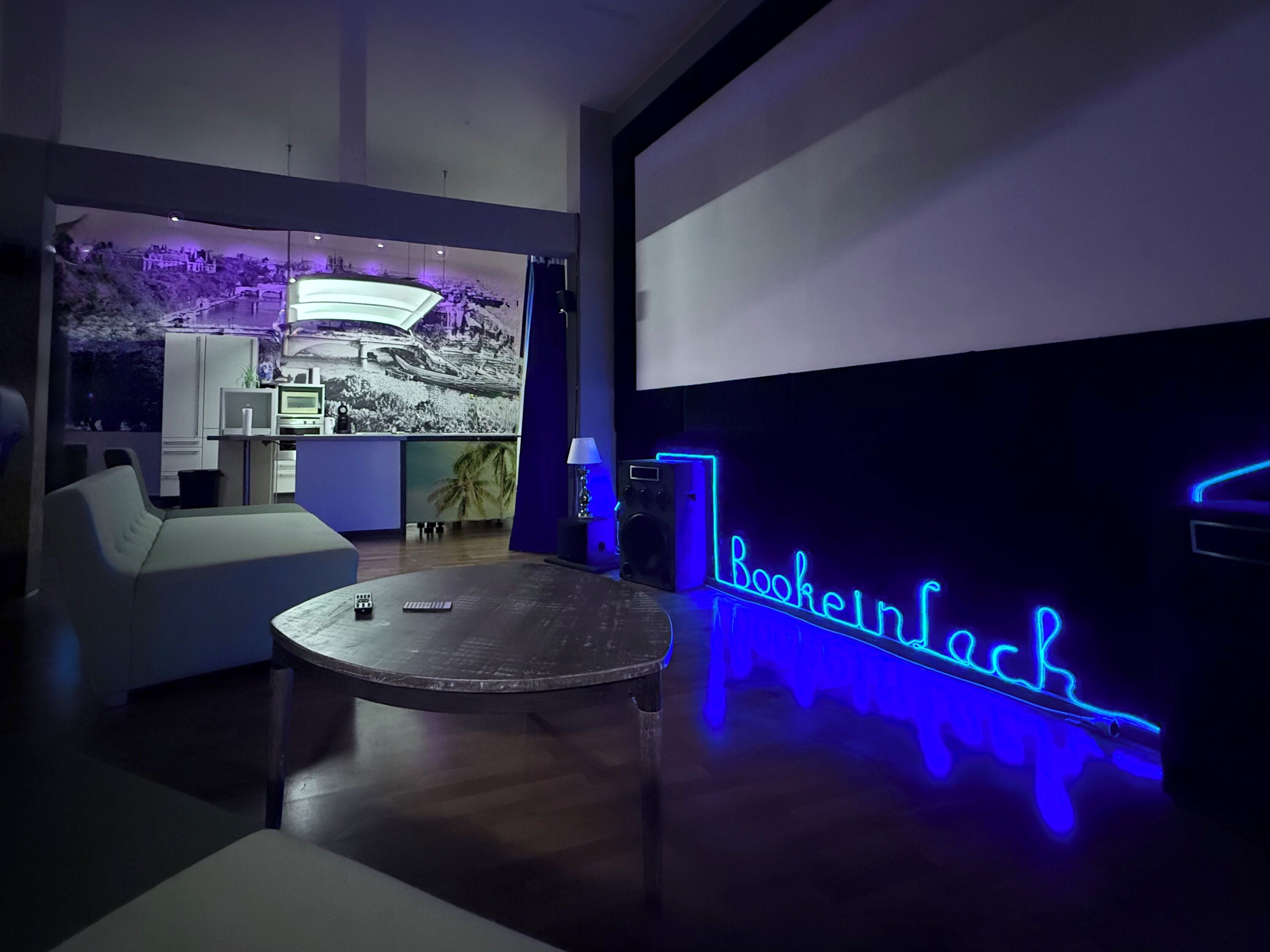 A modern lounge area with a neon sign reading "Bookenack," a comfortable seating area, and a projection screen displaying a landscape image.