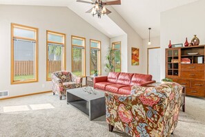 Ein geräumiges Wohnzimmer mit hohen Decken, großen Fenstern, einem roten Ledersofa, floralen Ohrensesseln, einem Couchtisch und einer Holzkabine mit Dekorationsgegenständen.